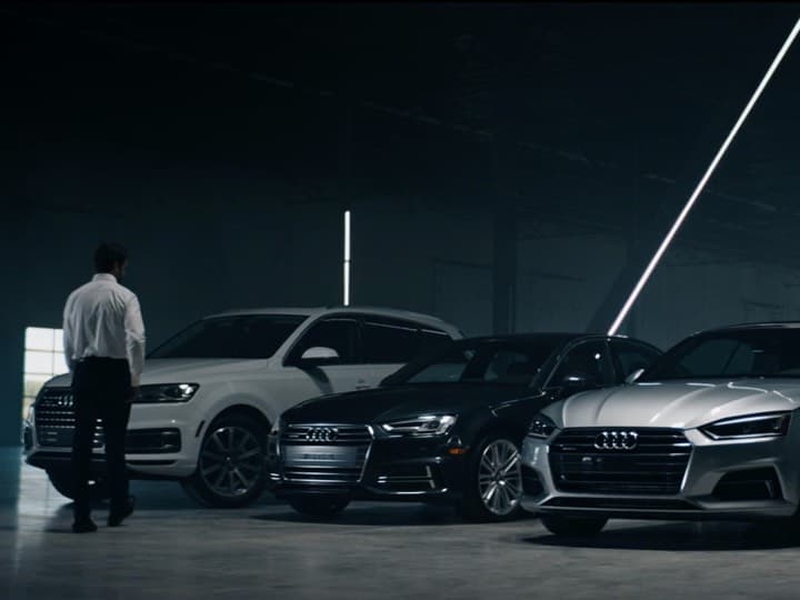 Concierge approaching a lineup of Audi vehicles including a white Q7, dark A4, and silver A5 in a dramatic showroom